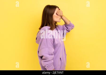 Don't want to look. Side view portrait of woman keeping hand on eyes and avoiding watch at something shameful, gross, wearing purple hoodie. Indoor studio shot isolated on yellow background. Stock Photo