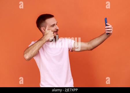 Man talking on video call and gesturing thumbs up, like sign, having online conversation on mobile phone, taking selfie, wearing pink T-shirt. Indoor studio shot isolated on orange background. Stock Photo