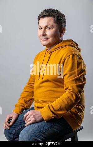 Portrait of attractive middle aged man looking at camera, sitting on chair and expressing positive emotions, wearing urban style hoodie. Indoor studio shot isolated on gray background. Stock Photo
