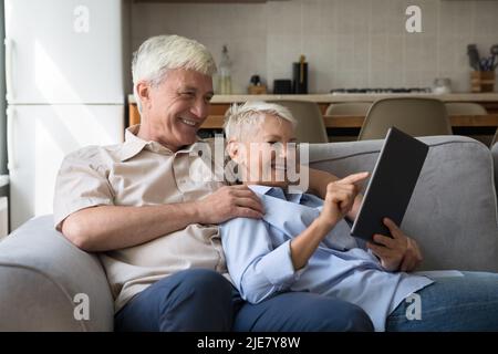 Happy older spouses relax on sofa using digital tablet Stock Photo