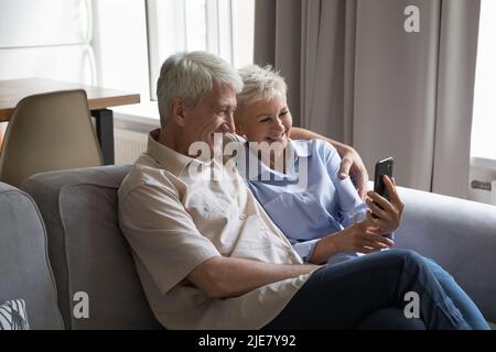 Happy mature couple sit on sofa using modern smartphone Stock Photo
