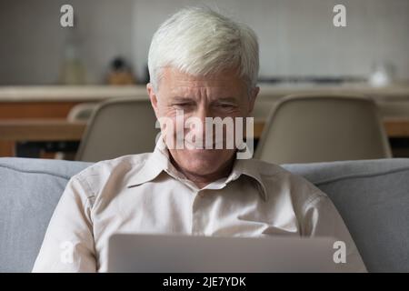 Smiling senior man sit on sofa using laptop Stock Photo