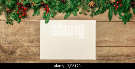 Christmas rustic mockup. sheet of white paper lies on an old table decorated with Christmas fir branches, red berries and cones. Christmas tree branch Stock Photo