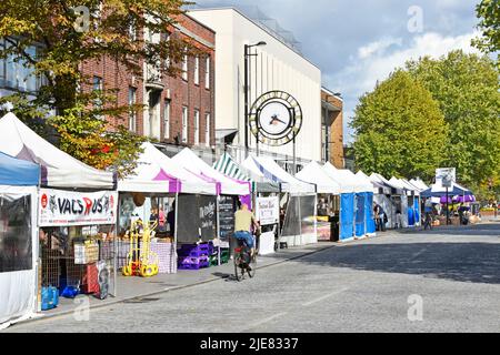Shopping high street view pavement roadside market stalls in Brentwood Essex cyclists pedalling along cobble stone road clock wrong time England UK Stock Photo