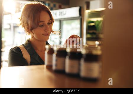 Happy young woman reading the label of a food product in the jarred food section of a grocery store. Cheerful young woman grocery shopping in a trendy Stock Photo