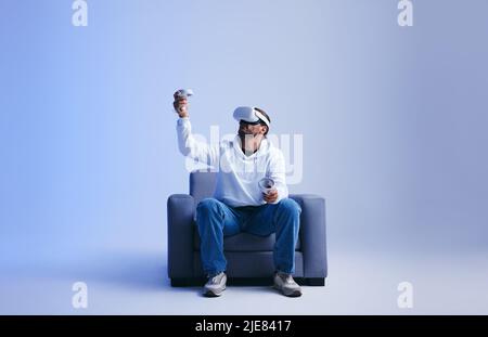 Young man playing an immersive virtual reality game on a couch. Man gaming with virtual reality goggles and controllers. Young man experiencing a 3D s Stock Photo