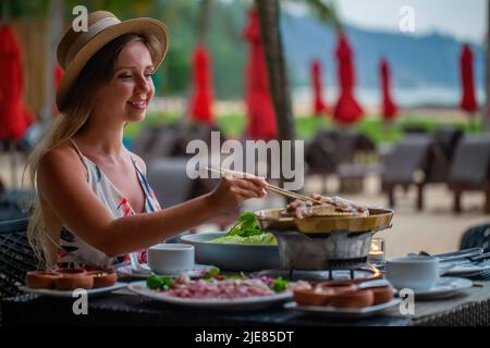 Happy smiling travel woman roasted raw pork slice on Korean barbecue pan. Female traveler cooking Thai style bbq, enjoy eating, grilling pork meat on Stock Photo