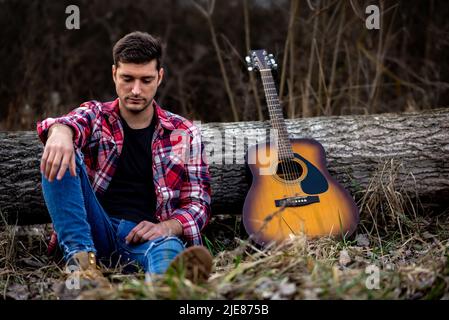 A handsome young man is sitting on ground leaning on a fallen tree next to an acoustic guitar in a nature park. Stock Photo