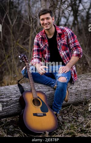 A handsome young man is sitting on a fallen tree next to an acoustic guitar in a nature park. Stock Photo