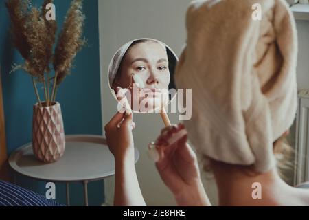 Close up mirror, woman using jade facial roller for face massage sitting on bed in bedroom looking in the mirror Stock Photo