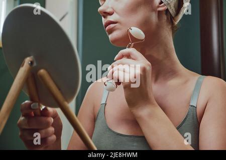 Close up woman using jade facial roller for face massage sitting by the bathroom mirror Stock Photo