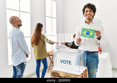 Young brazilian voter man smiling happy holding brazil flag standing by ballot at vote center. Stock Photo