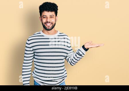 Young arab man with beard wearing casual striped sweater smiling cheerful presenting and pointing with palm of hand looking at the camera. Stock Photo