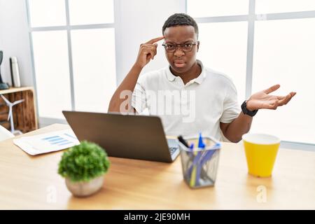 Young african man working at the office using computer laptop confused and annoyed with open palm showing copy space and pointing finger to forehead. Stock Photo