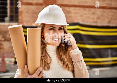 Young blonde woman holding blueprints talking on the smartphone at street Stock Photo