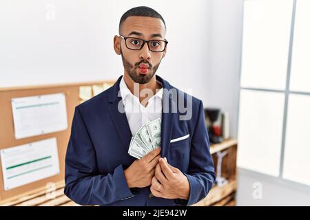 African american young man hiding dollars in jacket making fish face with mouth and squinting eyes, crazy and comical. Stock Photo