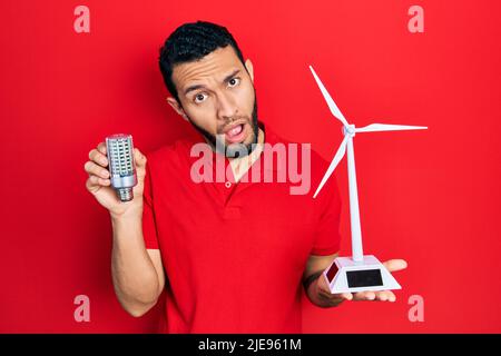 Hispanic man with beard holding solar windmill for renewable electricity and led bulb in shock face, looking skeptical and sarcastic, surprised with o Stock Photo