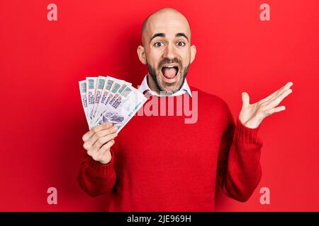 Young bald man holding egyptian pounds banknotes celebrating achievement with happy smile and winner expression with raised hand Stock Photo