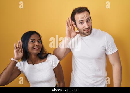 Interracial couple standing over yellow background smiling with hand over ear listening an hearing to rumor or gossip. deafness concept. Stock Photo