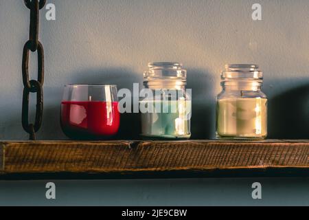 three colorful scented candles on a rustic wall shelf made from old reclaimed barn wood with rusty chain Stock Photo