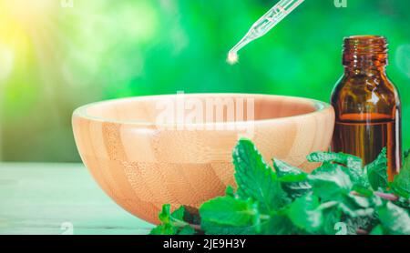 The peppermint extract in small bottle near peppermint leaf on wooden table. The essential oil falling from glass dropper into organic bio alternative Stock Photo