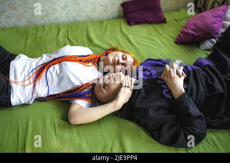 Portrait of a young tattooed punk couple of a girl and a guy with long dyed hair braided, lying on a large bed. Hugging each other. Stylish modern you Stock Photo