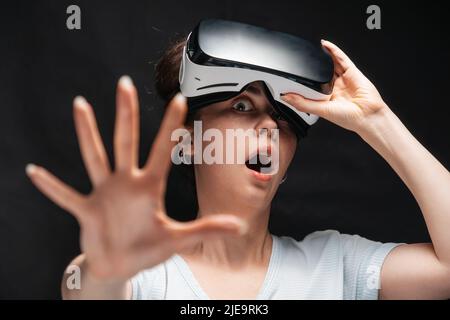 Portrait of young had shocked screaming woman removing VR glasses and reches hand. Black background. The concept of metaverse and cyberspace. Stock Photo