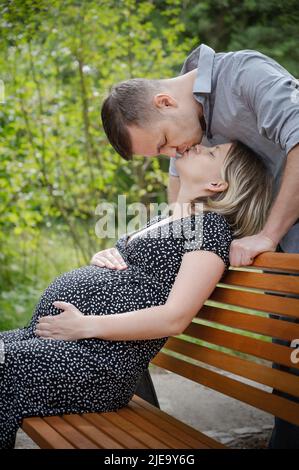 Pregnant woman with husband on bench in public park. Man kissing his wife. Loving couple enjoying together - waiting birth of baby. Stock Photo