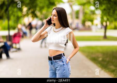 Portrait of young attractive dark-haired woman speaking on mobile phone at summer green park. Stock Photo