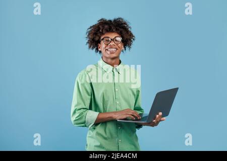 Positive black teen guy using his new modern laptop, typing on keyboard and smiling at camera, blue studio background Stock Photo