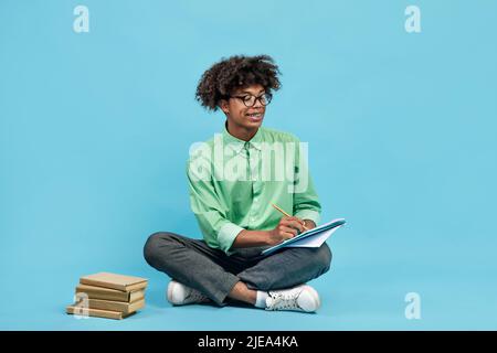 Smart black teen guy in glasses taking notes, holding notepad and pen, sitting with books nearby on blue background Stock Photo