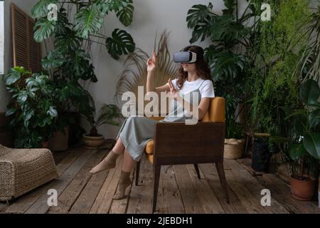 Impressed woman using virtual reality glasses touching air, resting in cozy tropical indoor garden Stock Photo