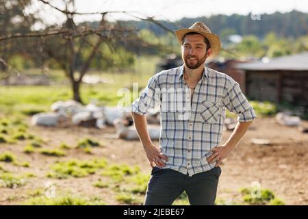 Portrait of young Caucasian handsome bearded male farmer smiling to camera. Happy rancherman in straw hat at the eco farm. Industrial production of Stock Photo