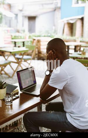 Vertical rearview confused african afro american businessman with head in hands, use laptop in cafe. Video call and read Stock Photo