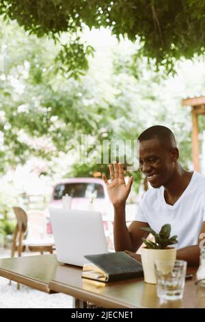 Smiling and glad multiracial man with laptop, raising arm, greet someone, looking at monitor. E-learning webinar meeting Stock Photo