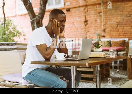 Puzzled and confused multicultural dark skin man using laptop, sit in cafe outdoors, looking at monitor. Job interview  Stock Photo
