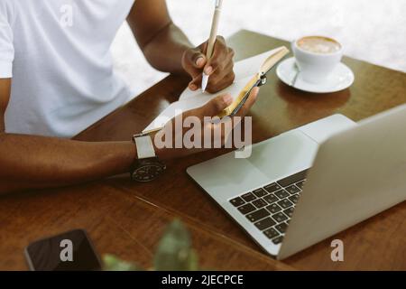 Unrecognizable cropped multicultural man using laptop, taking notes in workbook, drink coffee during internship study Stock Photo