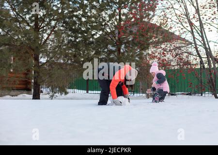 Nice small girl with mother rolling snow balls for building snowman on cottage backyard in evening with rowan and fir trees in background. Parents Stock Photo
