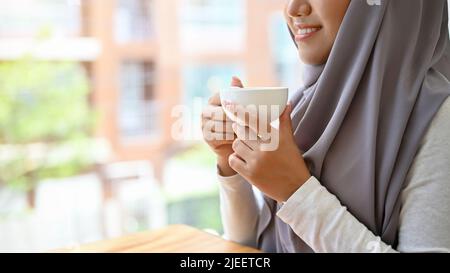 Beautiful attractive muslim woman with hijab relaxes sipping a morning coffee at home or office. cropped image, hands holding a coffee cup. Stock Photo