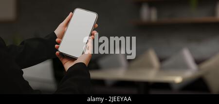 A female hands holding a mobile phone white screen mockup over blurred modern comfortable living room with comfortable sofa in the background. Stock Photo