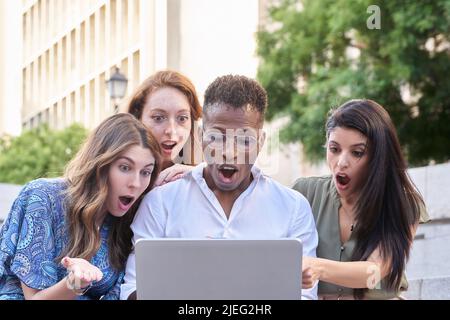 Group of multiracial people looking shocked at the laptop screen while using it outdoors in the street. Urban lifestyle, technology and friendship concept. Stock Photo