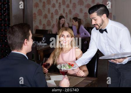 Polite waiter bringing ordered dishes to smiling couple at restaurant Stock Photo