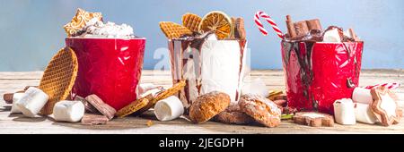 Christmas styled overloaded hot chocolate mugs with various toppings – chocolate slices, spices, sweets, cookies, candy and gingerbreads, wooden cozy Stock Photo