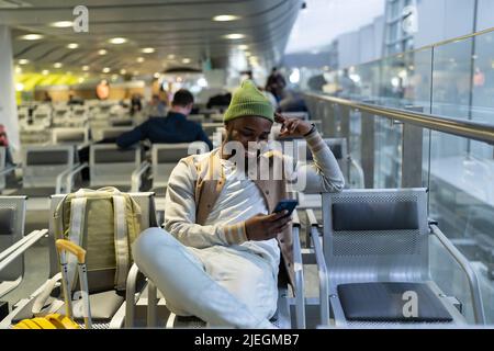 Happy hipster black man sitting in airport terminal using social media in smartphone waiting landing Stock Photo