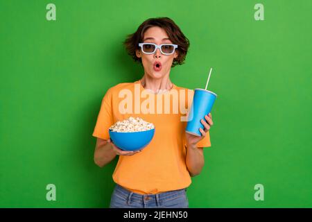 Photo of amazed funky young woman dressed orange t-shirt sunglass drinking beverage eating pop corn isolated green color background Stock Photo