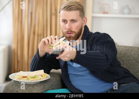 man on the sofa eating a sandwich pressing remote control Stock Photo