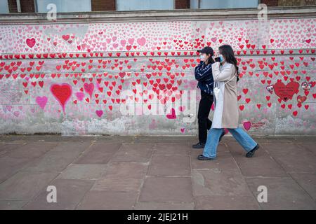 Two women wearing face masks walking in front of the National Covid memorial wall on the South Bank of the Thames, London UK.