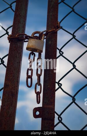 Close-up of a locked rusty old gate tethered by a chain and padlock with the blue sky and clouds blurred in the background. Concept of prison or loss Stock Photo