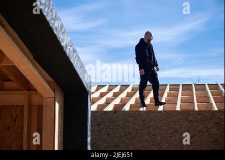 Male architect building wooden frame house. Bald man standing on construction site, inspecting quality of work on sunny day with blue sky on background. Stock Photo