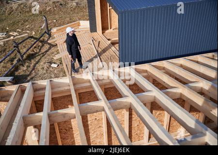 Male engineer building wooden frame house. Man developer on construction site, inspecting quality of work on sunny day, holding hammer in hands. Stock Photo